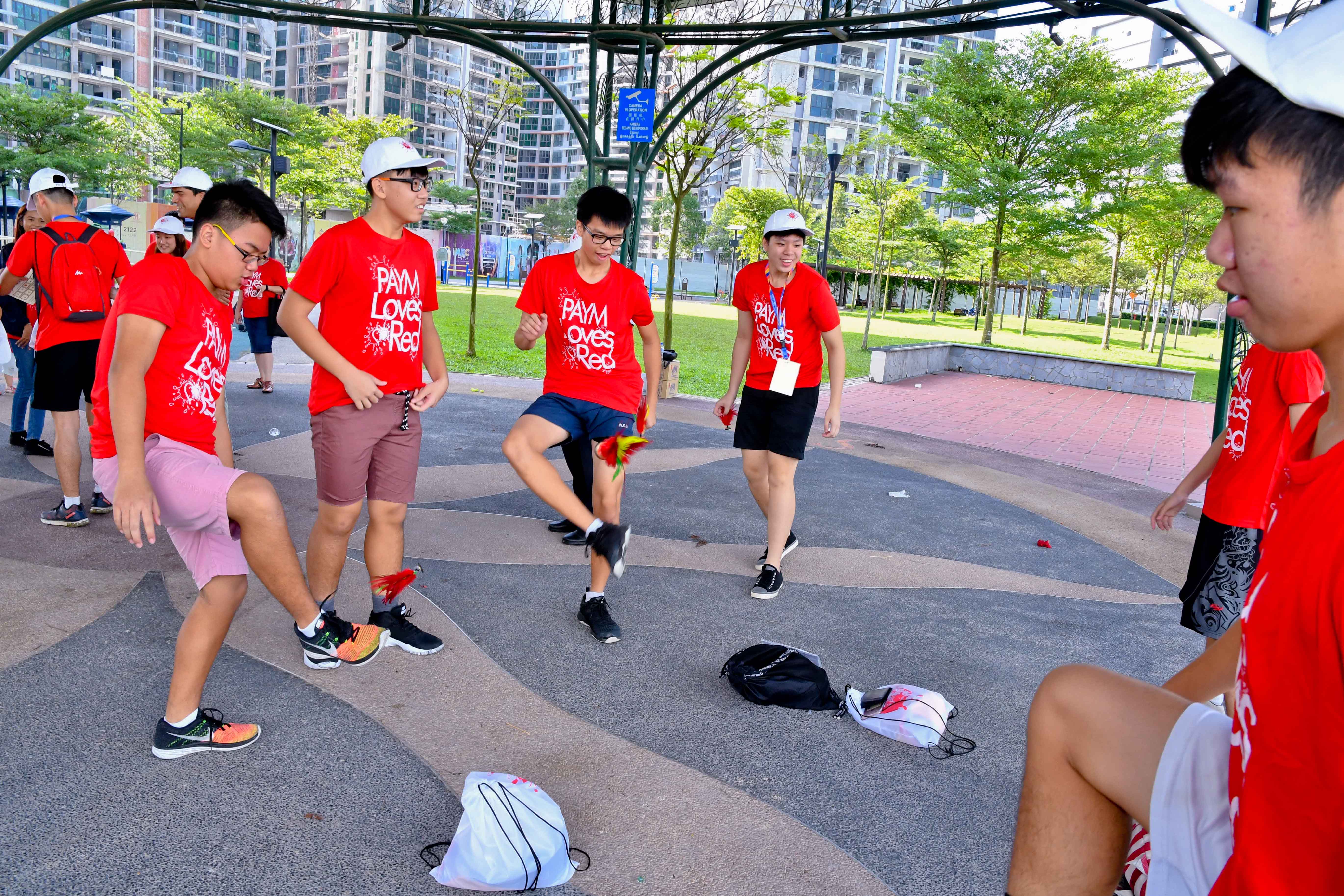 Several people in red "PAYM Loves Red" shirts are playing a game in a park with bags on the ground.
